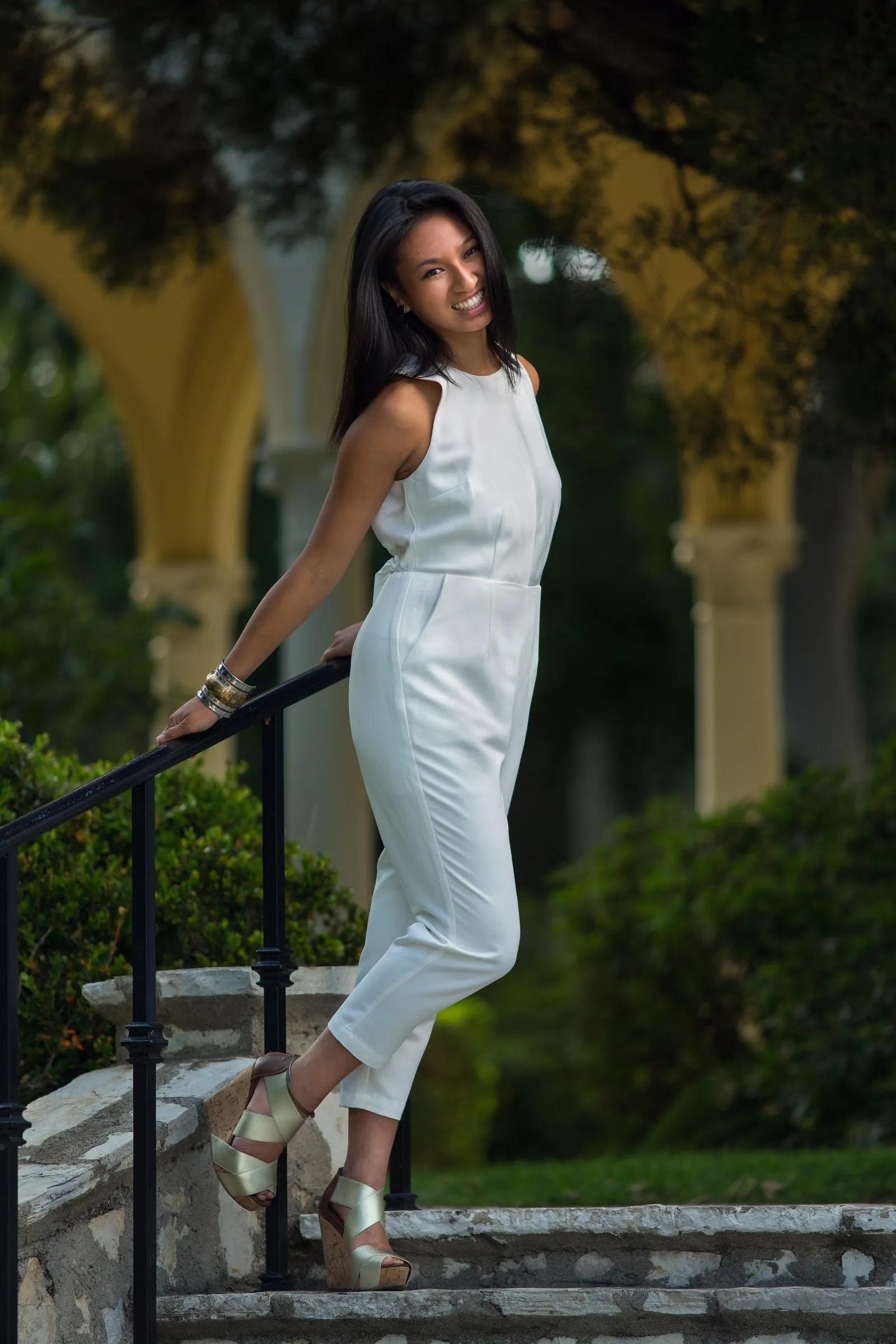 Gab smiling on stone stairs in a white jumpsuit, yellow arches behind her