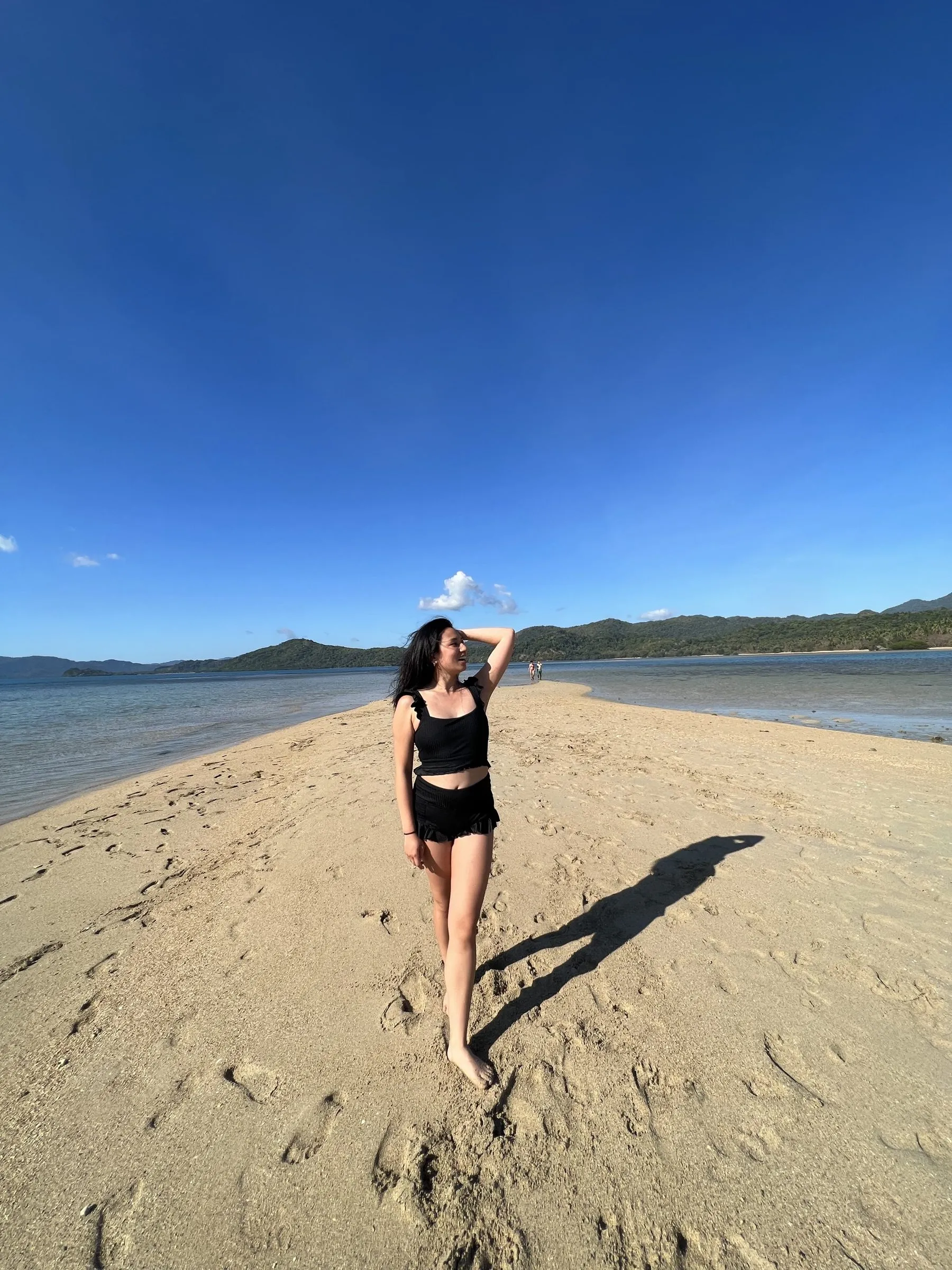 Gab standing on a sunlit sandbar between ocean and hills